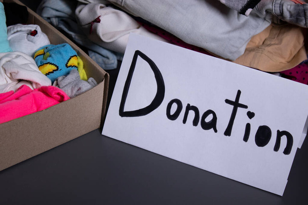 donation sign handwritten with black letters. A box with clothes and a pile of clothes nearby on a grey table.clothes donation concept. copy space - Photo, Image