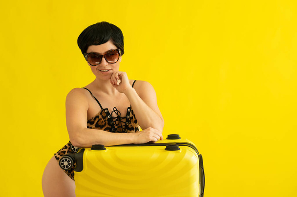 Portrait of an attractive woman in a one-piece leopard swimsuit and sunglasses with a suitcase on a yellow background. Seductive brunette in a monokini is ready to travel to the sea. Summer vacation. - Фото, зображення