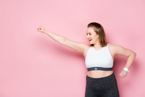 aufgeregtes übergewichtiges Mädchen zeigt Siegergeste, während es auf rosa wegschaut - Foto, Bild