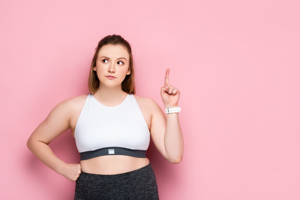 thoughtful overweight girl showing idea sign while holding hand on hip on pink - Фото, изображение
