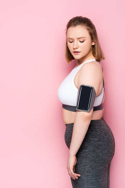 pretty overweight girl looking at smartphone in armband while standing on pink - Zdjęcie, obraz