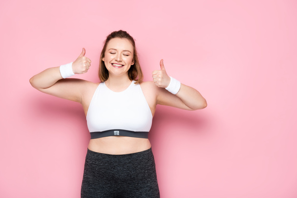 excited overweight girl with closed eyes showing thumbs up on pink - Φωτογραφία, εικόνα