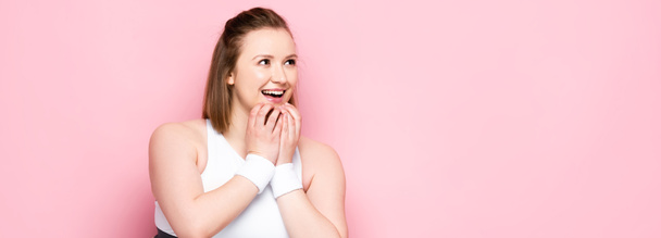 panoramic crop of excited overweight girl touching chin and looking away on pink - Photo, Image