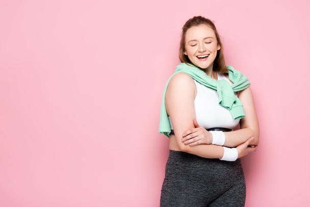 lächelndes übergewichtiges Mädchen mit Sweatshirt über den Schultern, stehend mit verschränkten Armen und geschlossenen Augen auf rosa - Foto, Bild