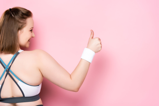 achteraanzicht van overgewicht meisje tonen duim omhoog op roze - Foto, afbeelding
