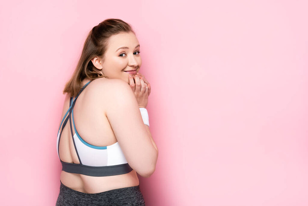 fröhliches übergewichtiges Mädchen berührt Kinn, während es in die Kamera auf rosa schaut - Foto, Bild