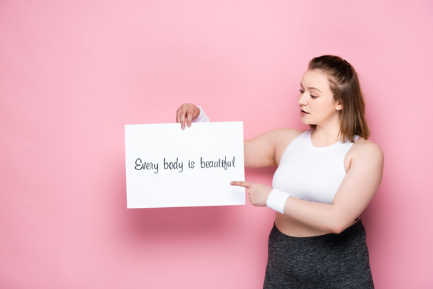 pretty overweight pointing at placard with every body is beautiful inscription on pink - Fotoğraf, Görsel