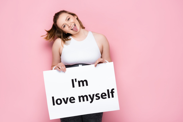 vrolijk overgewicht meisje houden plakkaat met ik hou van mezelf inscriptie en steken uit tong op roze  - Foto, afbeelding