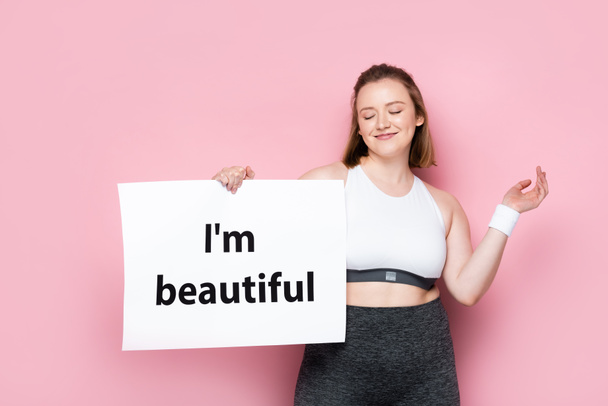 smiling overweight girl with closed eyes holding placard with i am beautiful inscription on pink - Photo, Image