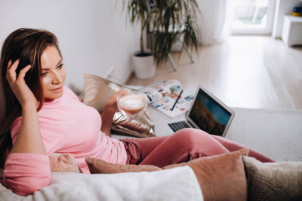 a girl in the morning in pajamas at home working on a laptop with drinking coffee, a girl self-isolated at home and resting on the couch and watching a laptop.Household chores. - Foto, imagen