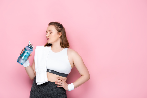confident overweight girl with towel on shoulder holding sports bottle while standing with hand on hip on pink - Foto, afbeelding