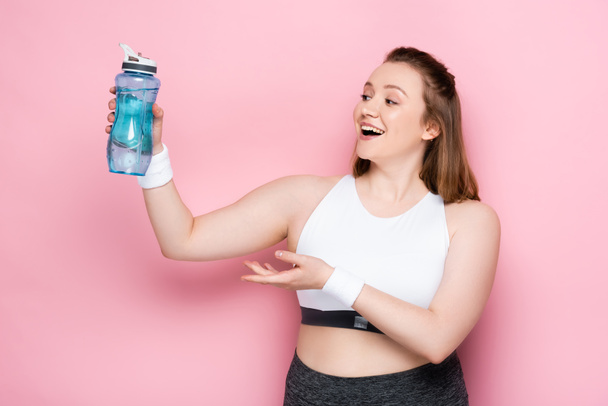 aufgeregtes übergewichtiges Mädchen mit Sportflasche in ausgestreckter Hand auf rosa - Foto, Bild