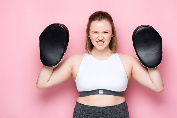 angry overweight girl in boxing pads grimacing while looking at camera on pink - Photo, Image