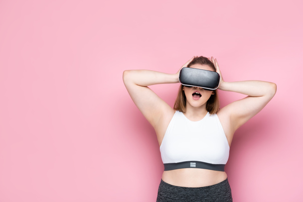 shocked size plus girl in sportswear and vr headset touching head on pink - Valokuva, kuva