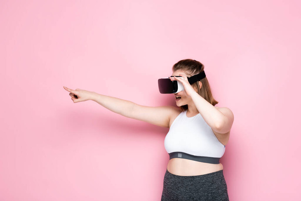 overweight girl in sportswear and vr headset pointing with finger on pink - Photo, Image