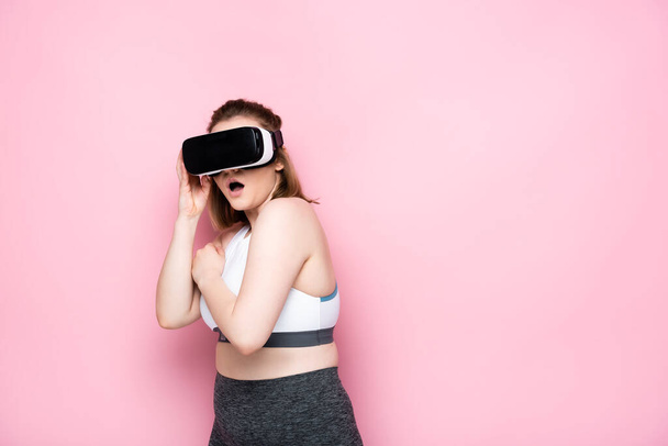 scared size plus girl in sportswear touching vr headset on pink - Photo, Image