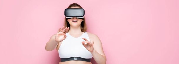 panoramic shot of smiling size plus girl in vr headset and sportswear smiling and gesturing on pink - Photo, Image