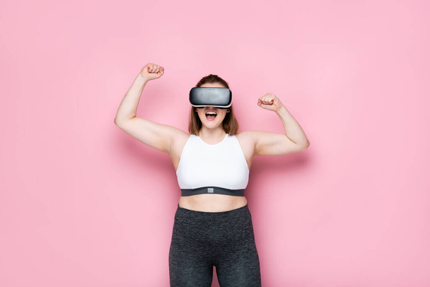 excited size plus girl in vr headset and sportswear showing winner gesture on pink - Foto, afbeelding