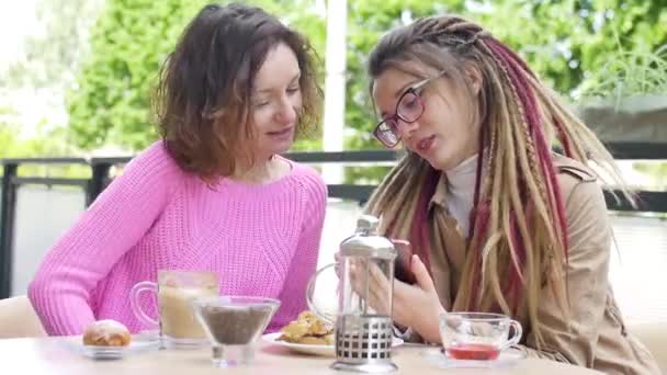 Modernes Mädchen mit langen Dreadlocks zeigt ihrer schönen Freundin in rosa Pullover während einer gemeinsamen Mittagspause in einem Café im Freien etwas auf dem Smartphone - Filmmaterial, Video