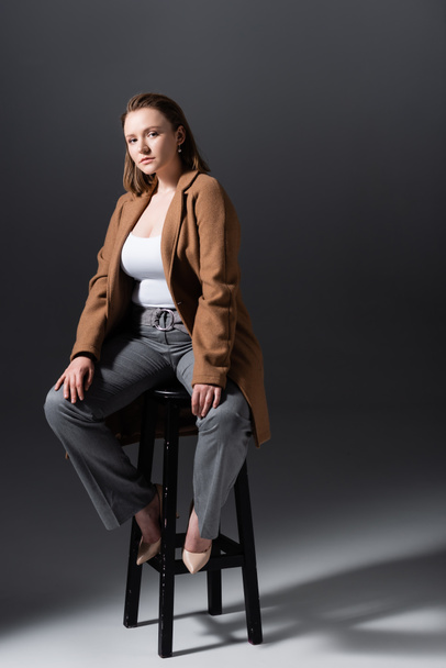 full length view of attractive, stylish overweight girl sitting on high stool and looking at camera while posing on grey - Foto, Imagem