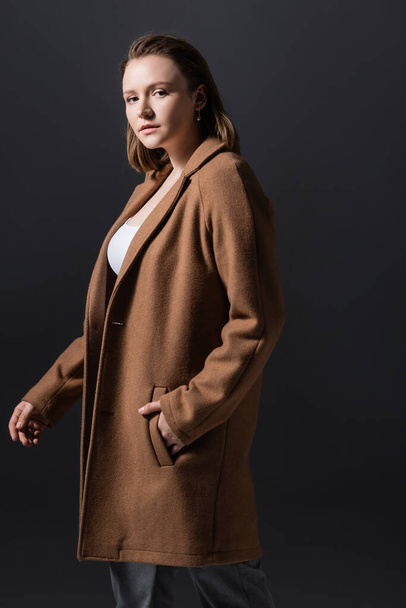 beautiful, confident overweight girl in coat looking at camera while holding hand in pocket isolated on grey - Valokuva, kuva