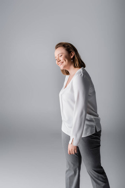 smiling, elegant overweight girl in white blouse posing with closed eyes on grey - Foto, Imagem