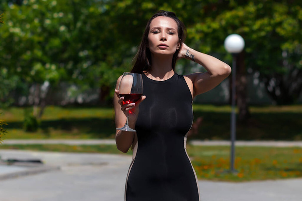 Ελκυστική κυρία σε μαύρο κομψό φόρεμα αγγίζοντας το λαιμό και απολαμβάνοντας κόκκινο κρασί την ηλιόλουστη μέρα του καλοκαιριού στο πάρκο - Φωτογραφία, εικόνα