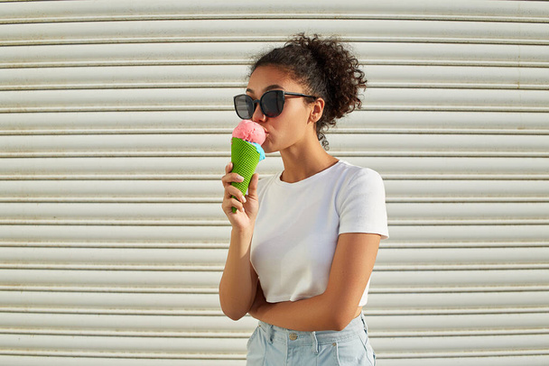 a young beautiful African-American girl in a white t-shirt and light jeans eats ice cream against a light wall on a Sunny day. - Valokuva, kuva