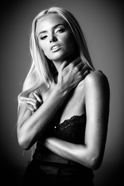 Porträt einer blonden jungen Frau mit wunderschönem Make-up und Frisur in einem schwarzen Spitzenkleid steht auf grauem Hintergrund. Modemodel posiert in eleganter Kleidung im Studio. Schwarz-weiß, monochrom - Foto, Bild