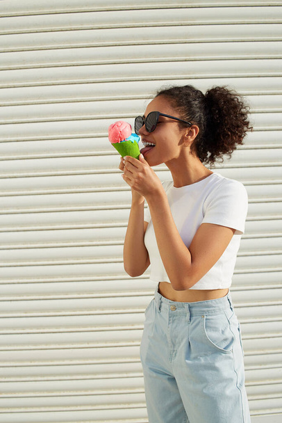 молодая красивая афроамериканка в белой футболке и легких джинсах ест мороженое у светлой стены в солнечный день
. - Фото, изображение