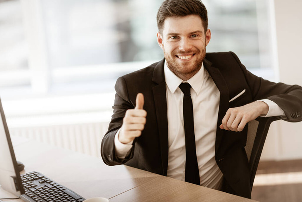 Business concept. Successful young businessman at work. Manager sitting at the office table happy showing thumb up. Man smiling in suit indoors on glass window background - Photo, Image
