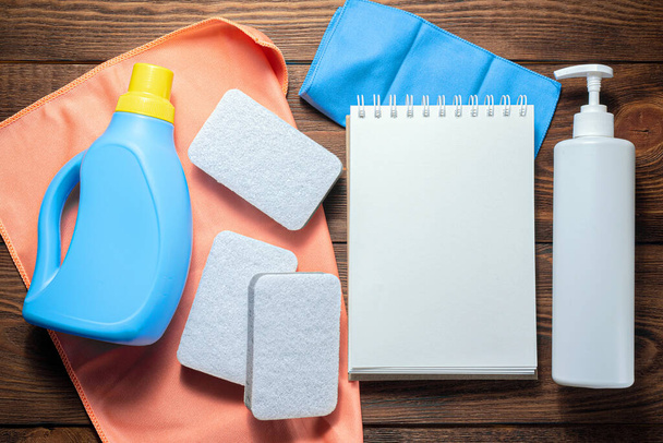 Washing supply, softener, detergent, dish cloth and blank page notepad on wooden table. Household tips mock up. - Φωτογραφία, εικόνα