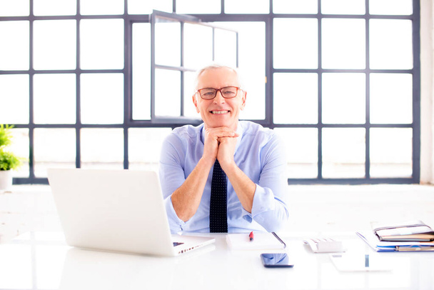 Portrait shot happy senior businessman wearing shirt and tie while sitting with folded arms behind his laptop in the office. - 写真・画像