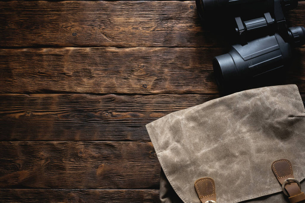Travel or adventure flat lay background with copy space. A backpack and a binoculars on a brown wooden table. - Фото, изображение