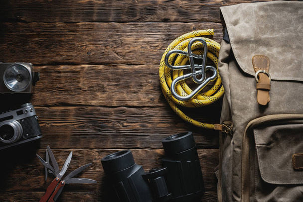 Travel or adventure flat lay background with copy space. A backpack, photo camera, flashlight, multitool knife, rope, carbines and a binoculars on a brown wooden table. - Fotoğraf, Görsel