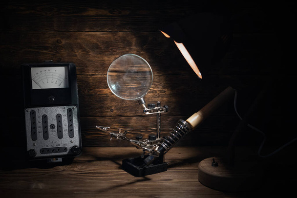 Retro analog voltmeter and soldering iron in the light of lamp on a brown wooden table background. Electrical works abstract background.  - Фото, изображение