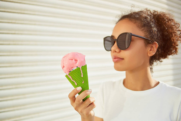una joven hermosa afroamericana con una camiseta blanca y jeans ligeros come helado en un día soleado. enfoque selectivo. área de enfoque pequeña
. - Foto, imagen