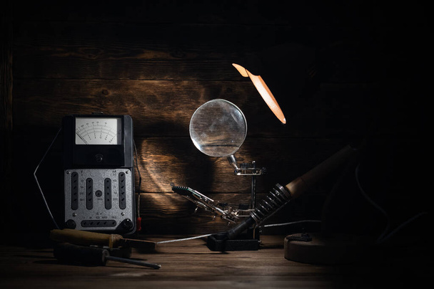 Retro analog voltmeter, soldering iron, pliers and screwdriver in the light of lamp on a brown wooden table background. Electrical works abstract background.  - Φωτογραφία, εικόνα