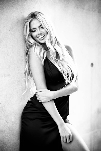 Blondynka młoda kobieta z pięknym makijażem i włosami w czarnej sukience stojącej w pobliżu szarej ściany. Modelka pozująca w eleganckich ciuchach na świeżym powietrzu w środowisku miejskim. Czarno-białe, monochromatyczne - Zdjęcie, obraz