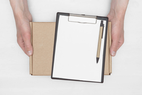 A parcel cardboard box in a delivery boy hands and a blank invoice document mock up on a white wooden table background. Delivery service concept. - 写真・画像