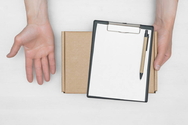 A parcel cardboard box in a delivery boy hands and a blank invoice document mock up on a white wooden table background. Delivery service concept. - Zdjęcie, obraz