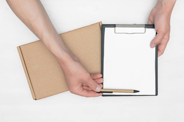 A parcel cardboard box in a delivery boy hands and a blank invoice document mock up on a white wooden table background. Delivery service concept. - Valokuva, kuva