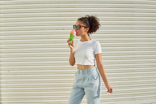 a young beautiful African-American girl in a white t-shirt and light jeans eats ice cream on a Sunny day. selective focusing. small focus area. - Foto, Imagem
