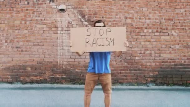 Maskeli beyaz adam elinde karton posterle kırmızı duvara karşı duruyor. Üzerinde "RACISM 'i durdurun" yazıyor. Tek protesto. - Video, Çekim