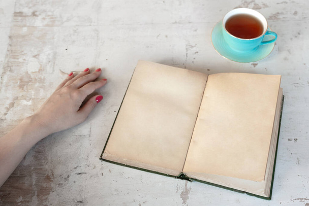 Woman is reading an open blank page book with a copy space and drinking a tea from a cup on a old white wooden table background. - Fotoğraf, Görsel