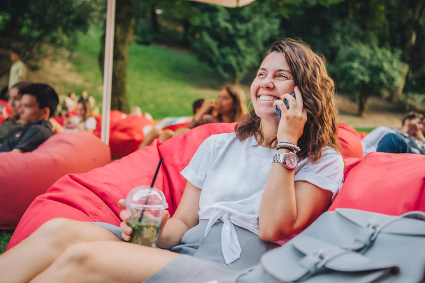 woman sitting outdoors drinking cocktail in public park surfing internet on the phone - Photo, Image