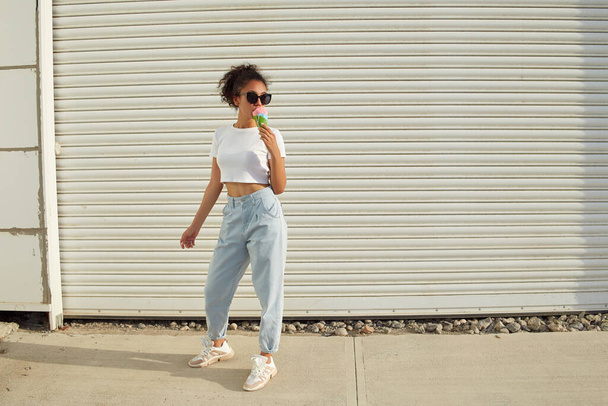a young beautiful African-American girl in a white t-shirt and light jeans eats ice cream on a Sunny day. selective focusing. small focus area. - Foto, Imagem