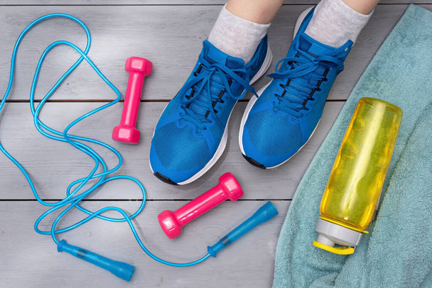 Sport gym background. Skipping rope, dumbbells, towel, bottle of water and a female feet in a blue sneakers on wooden floor background. - Photo, image