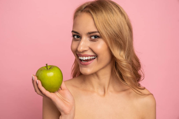 強い白いストレート歯のスタマトロジーのコンセプトをクリア。ピンクの背景に緑のリンゴを手に持つ魅力的なヌード自然モデルの肖像画. - 写真・画像