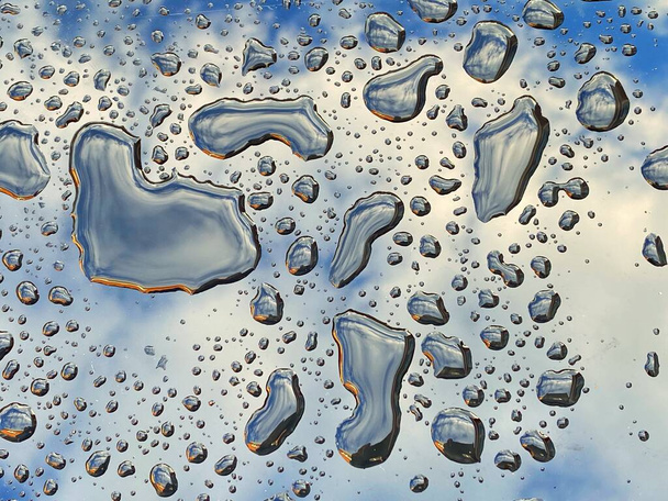 Vue rapprochée des globules d'eau sur une surface brillante après une pluie avec reflet du ciel bleu et des nuages - Photo, image